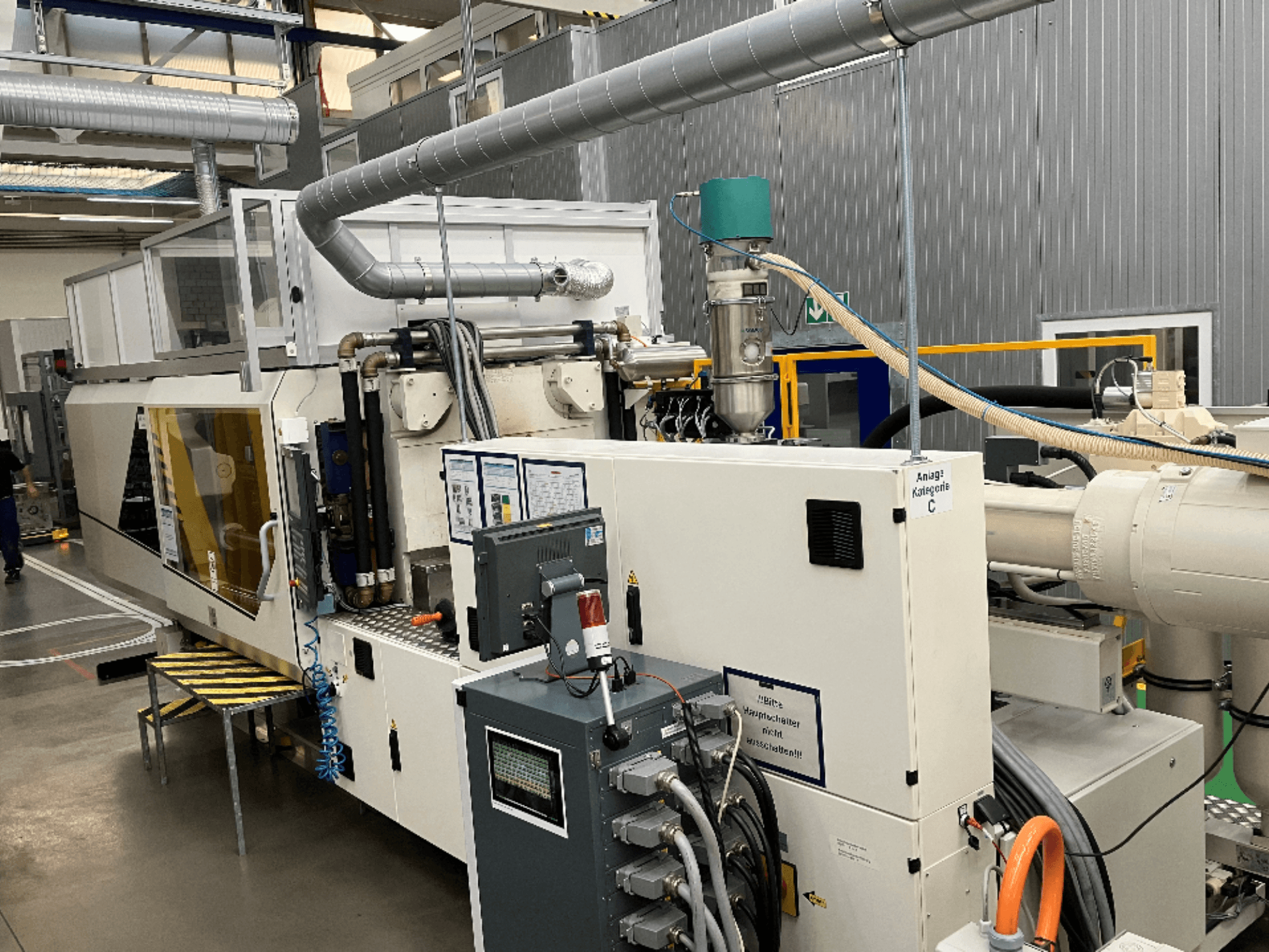 Vstřikovací stroj, pohled zepředu, se značkou, ovládacím panelem a viditelnými hadicemi v továrním prostředí.