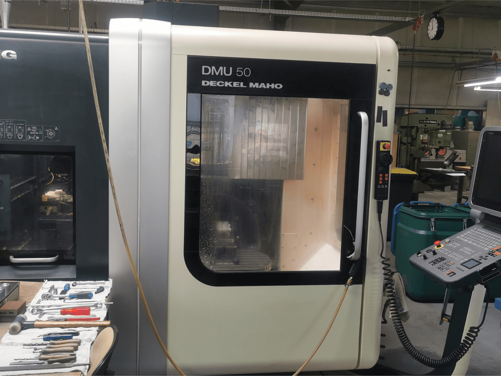 Stroj DMU 50 deckel maho CNC při pohledu zepředu, na kterém je vidět ovládací panel a průhledné dveře. Nástroje jsou poblíž.
