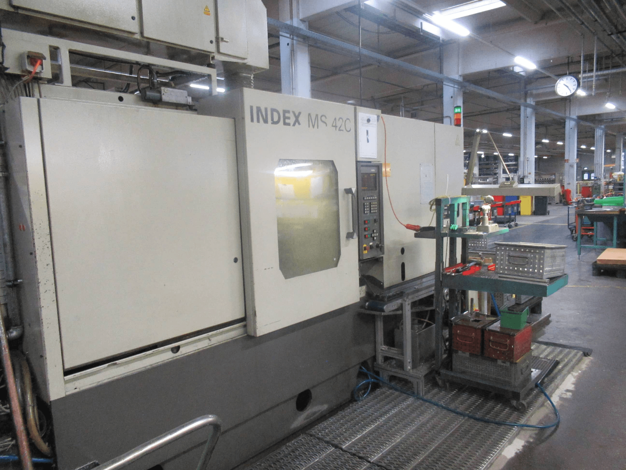 CNC stroj INDEX MS 42C v dílně, boční pohled ukazuje ovládací panel a úložný prostor pro nástroje na vozíku.