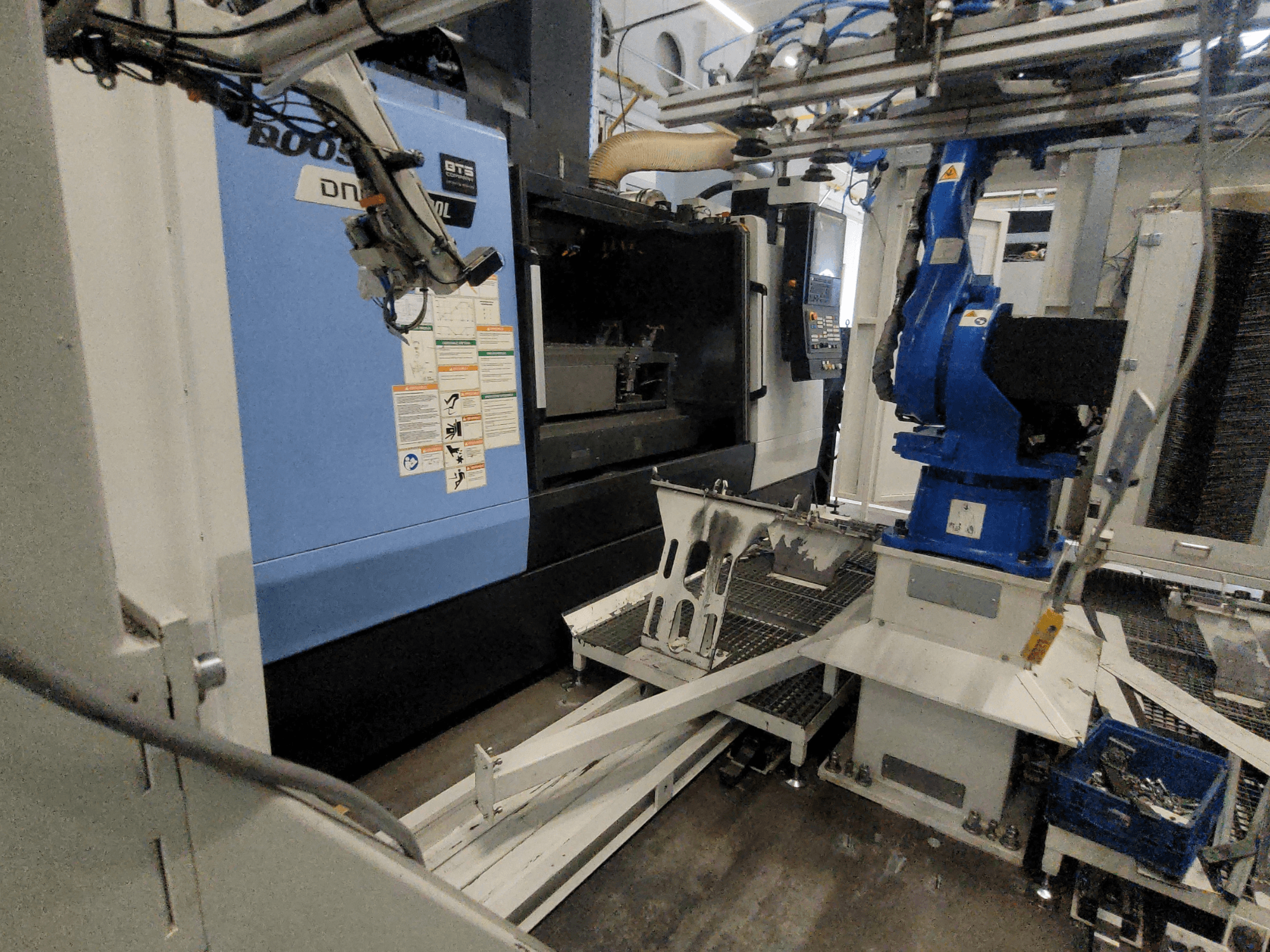 Boční pohled na vertikální obráběcí centrum DOOSAN DNM 6700L, na kterém je vidět robotické rameno a ovládací panel, znázorňující uspořádání stroje.