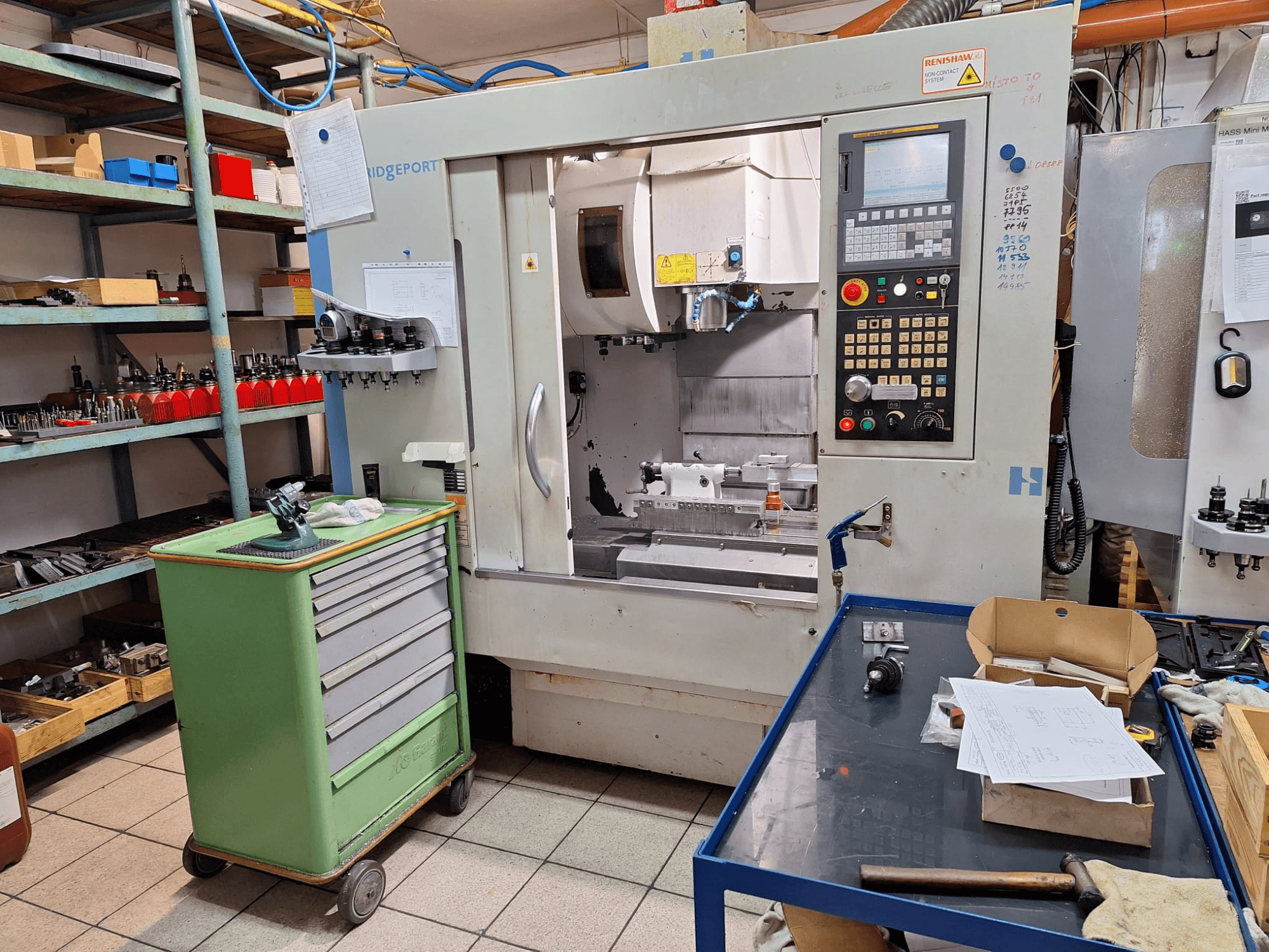 Pohled zepředu na vertikální obráběcí centrum Hardinge Bridgeport GX 710 s ovládacím panelem, nástroji a policemi v prostředí dílny.