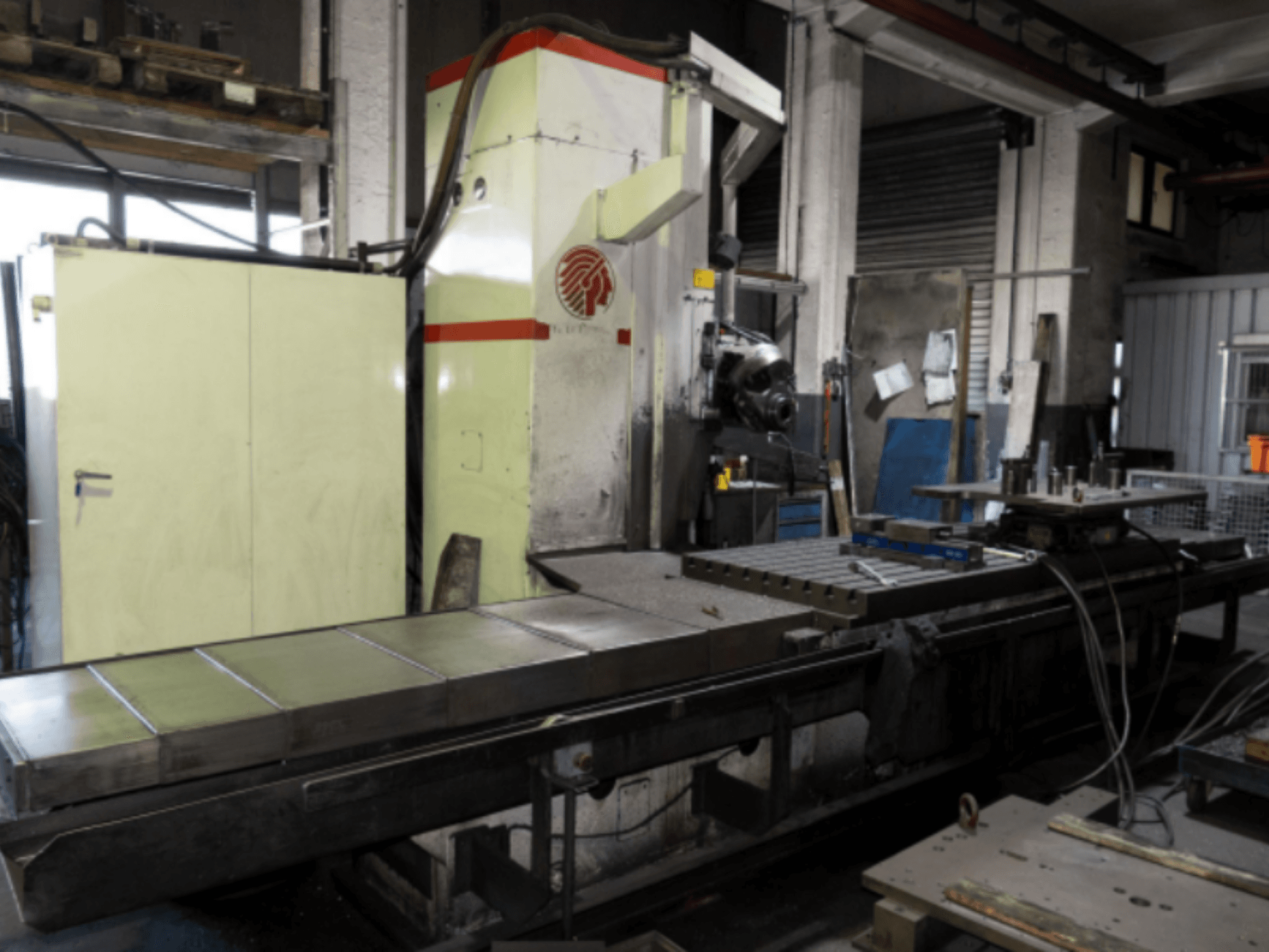 Průmyslová frézka s velkým stolem, boční pohled, ukázka ovládacích prvků a přídavných zařízení v dílenském prostředí.