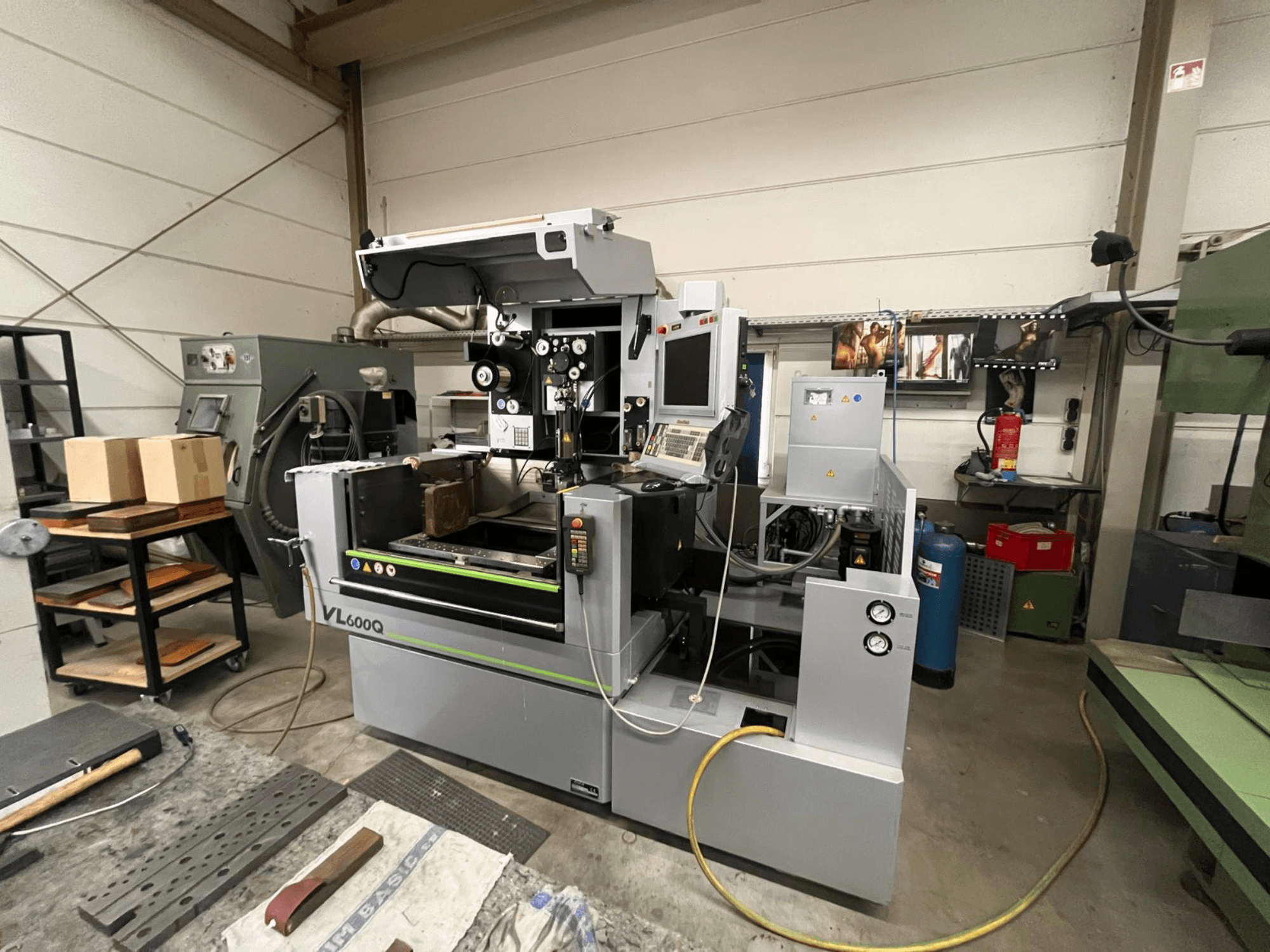 CNC stroj VL600Q v dílně, pohled zepředu, s ovládacím panelem, uloženým materiálem a různými nástroji v blízkosti.