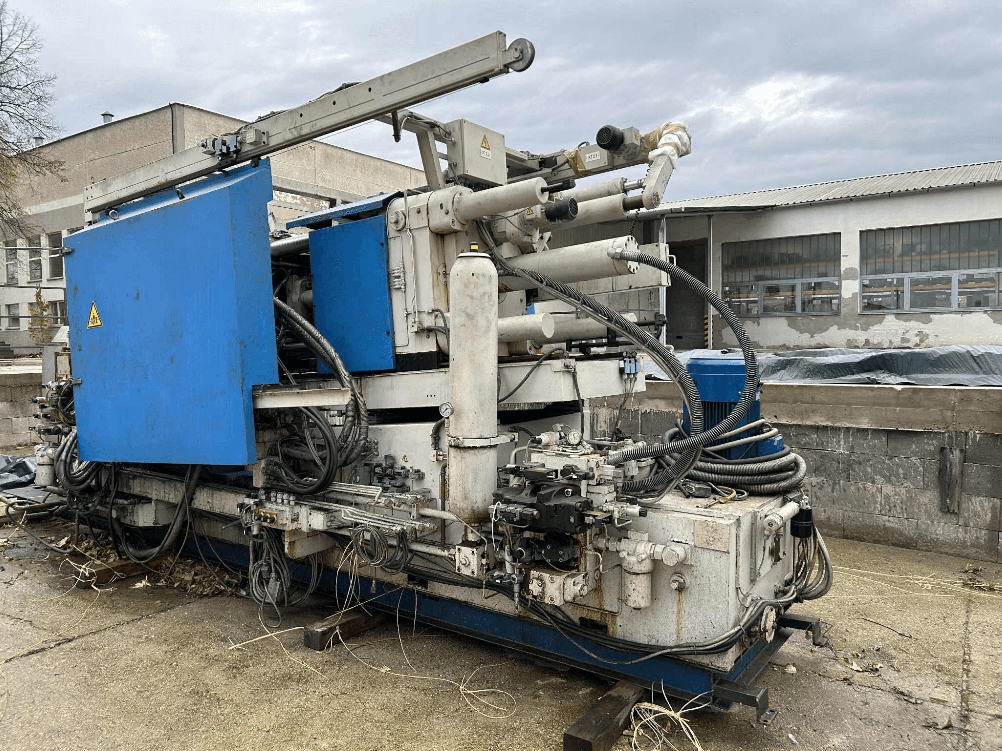 Velký průmyslový hydraulický stroj s modrým pláštěm, různými hadicemi a kovovými součástmi, pohled z boku.