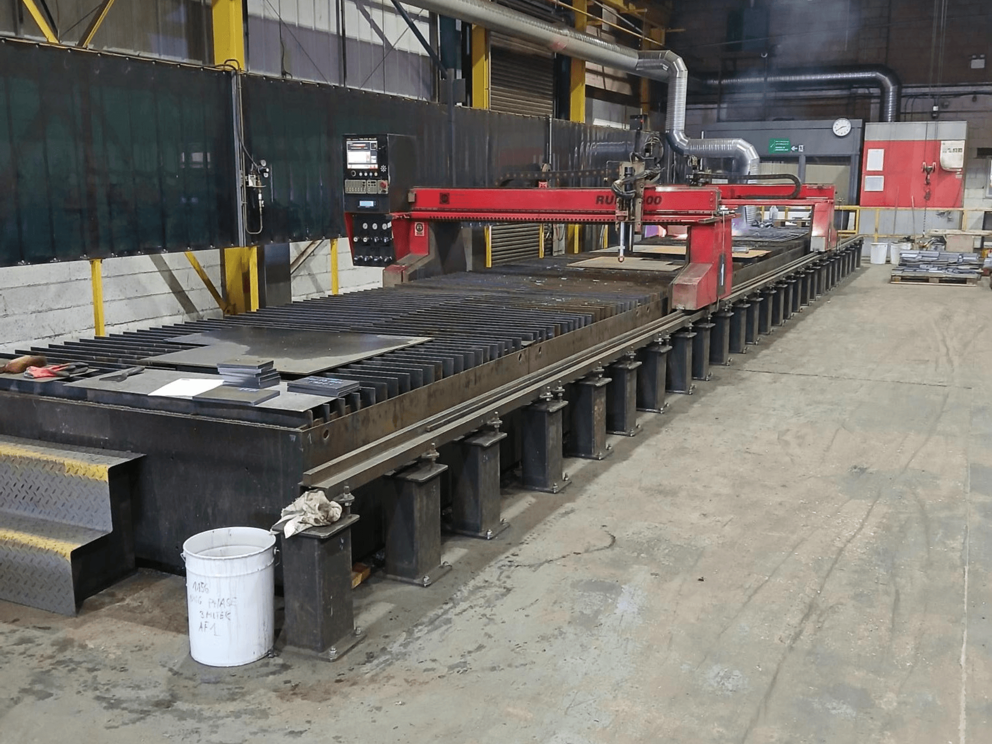 Červený řezací stroj RUIDA 300, boční pohled, s plechem na ocelovém stole, umístěný v průmyslovém pracovišti.