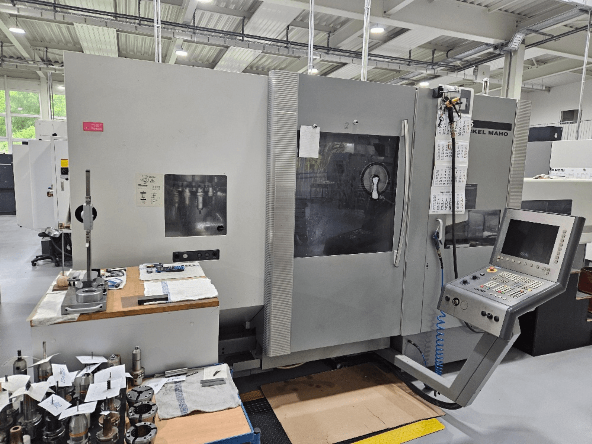 Pohled zepředu na stroj DMG Mori DMU 70 s ovládacím panelem, nástroji na dřevěném stole a různými obrobky v blízkosti.