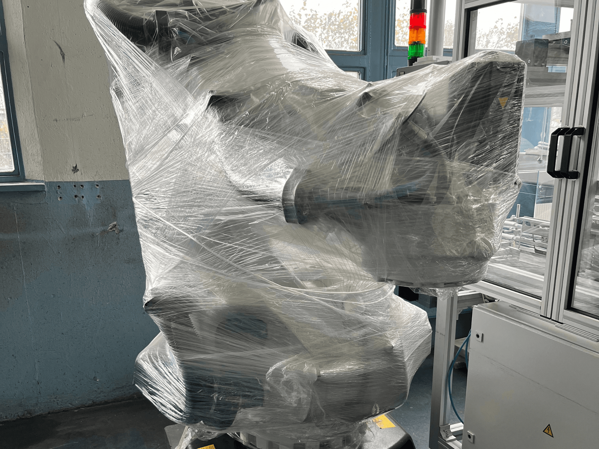 Průmyslové robotické rameno zabalené do plastu, pohled zepředu. Včetně řídicí jednotky a kontrolky. Značka není viditelná.