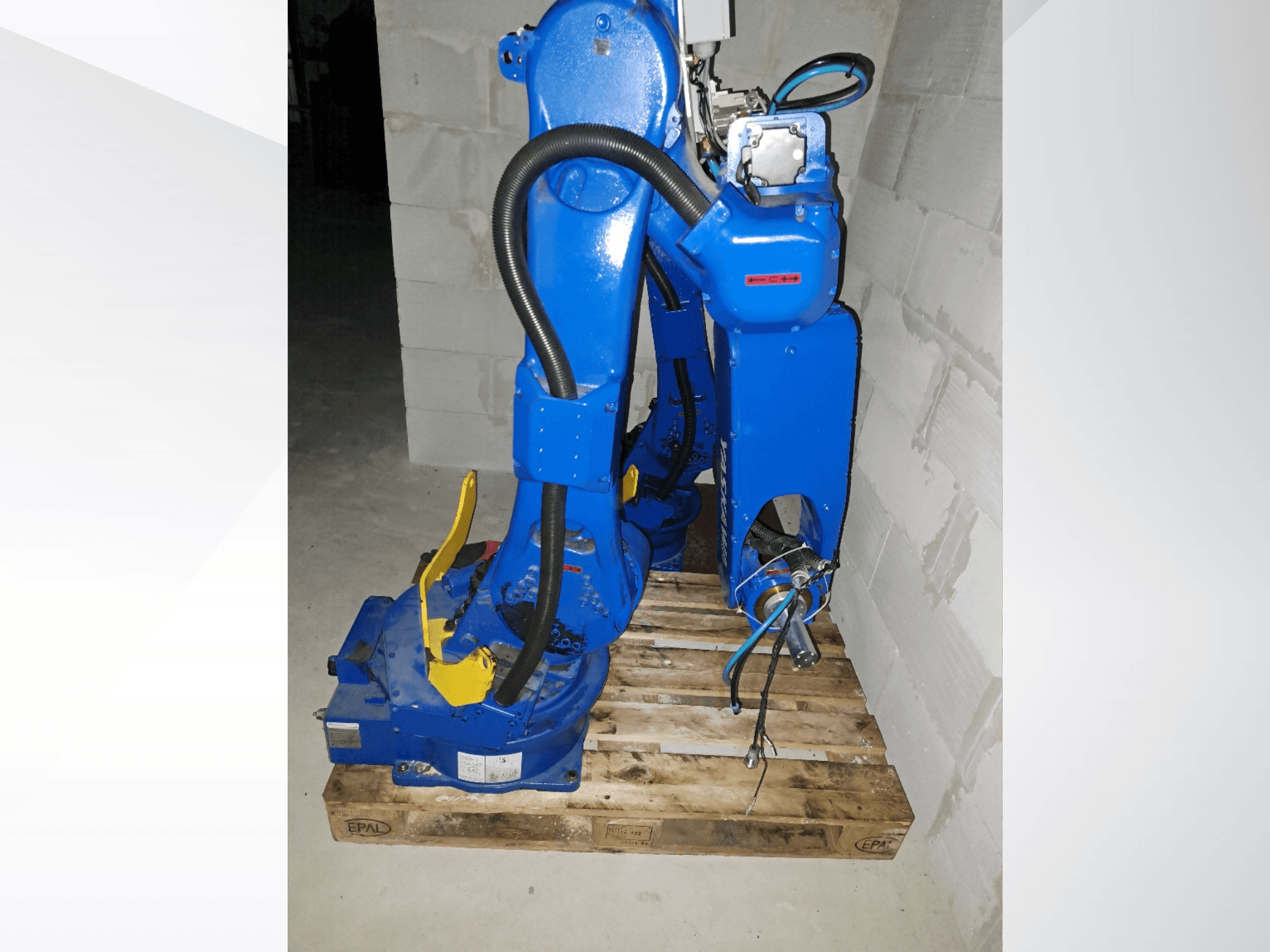 Robotické rameno Yaskawa Motoman GP25 v modré barvě, boční pohled na dřevěné paletě, s kabely a mechanickými klouby.