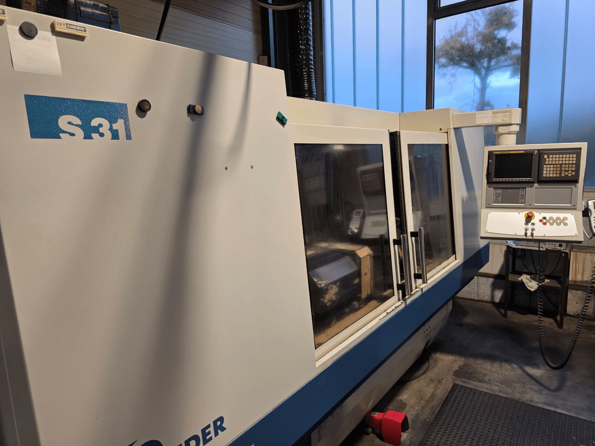 Bruska S31 CNC, pohled zepředu, ovládací panel s obrazovkou a tlačítky, velké skleněné dveře, průmyslové prostředí.
