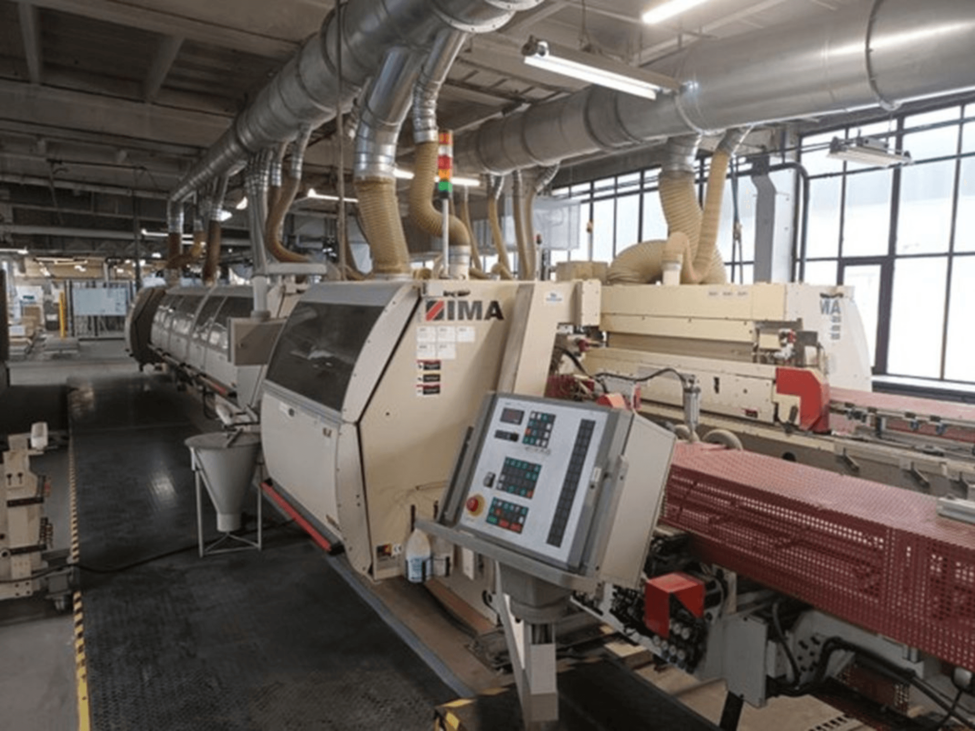 Průmyslové stroje IMA v továrním prostředí, pohled zepředu, s ovládacími panely a trubkami pro odsávání prachu.