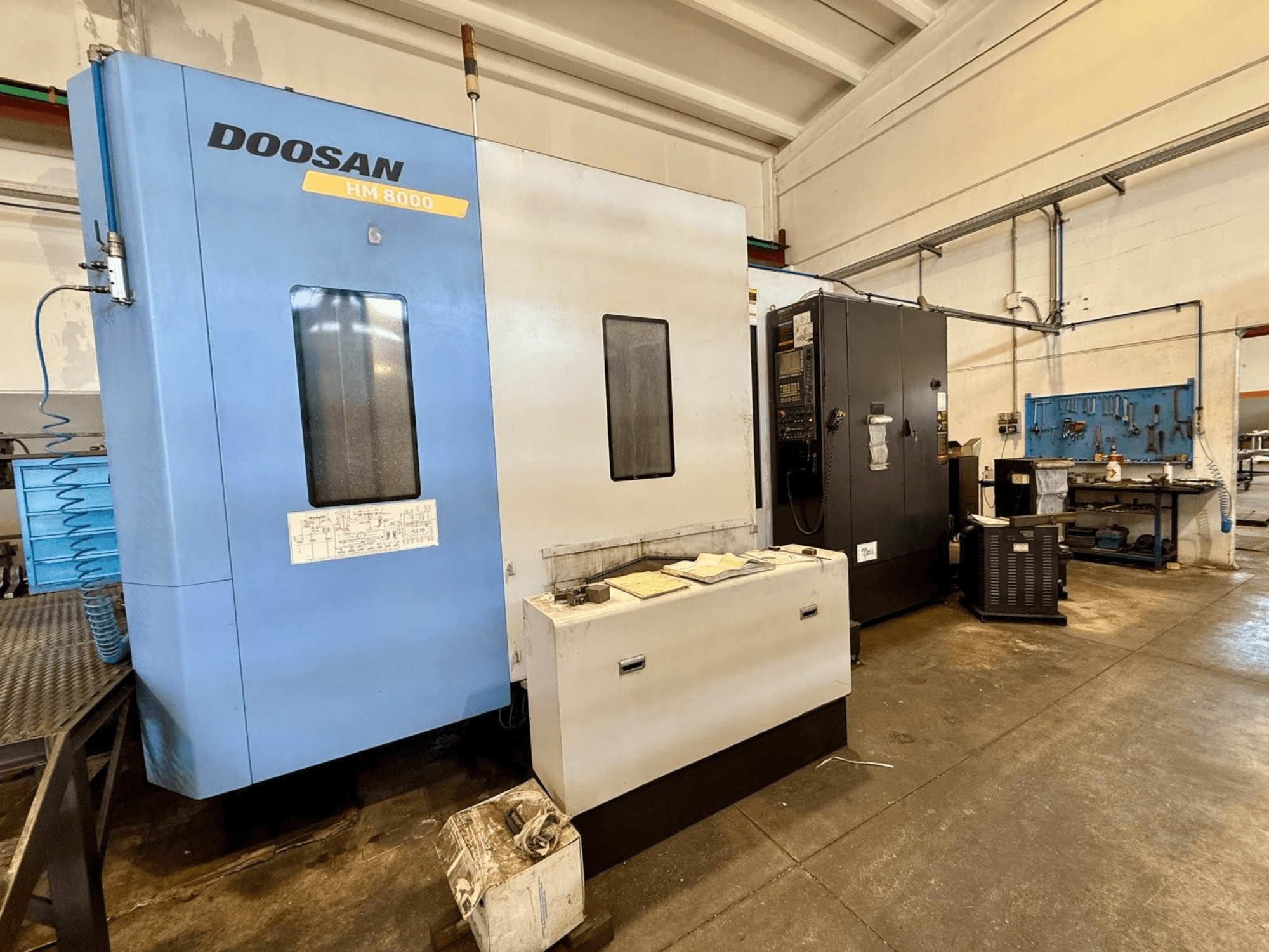 Horizontální obráběcí centrum DOOSAN HM 8000 v modré barvě, pohled zepředu, na kterém je vidět ovládací panel a okolní dílenské vybavení.