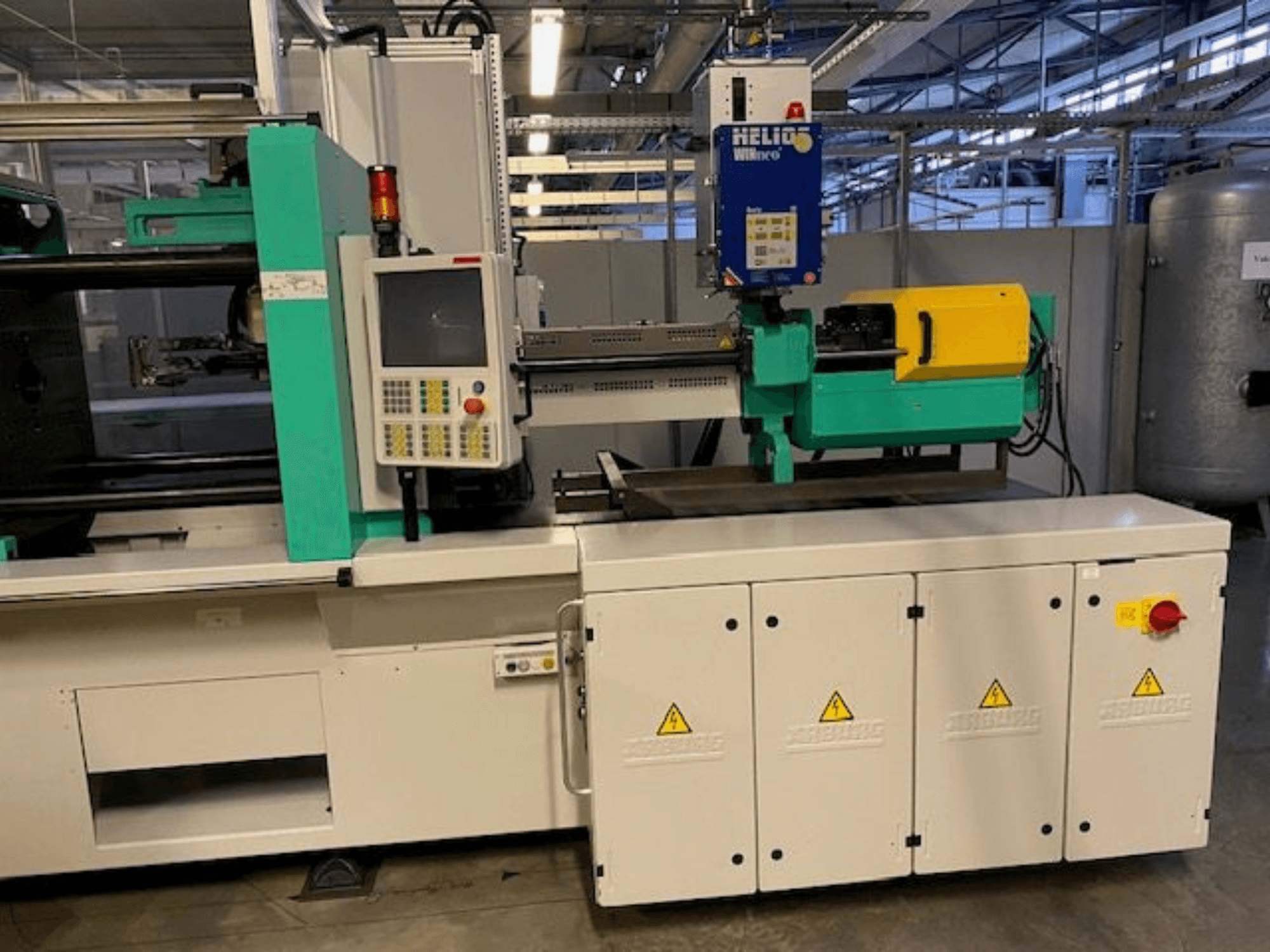 Průmyslový vstřikovací stroj, model HELIO, pohled zepředu, s ovládacím panelem, bezpečnostními kryty a tlačítkem nouzového zastavení.