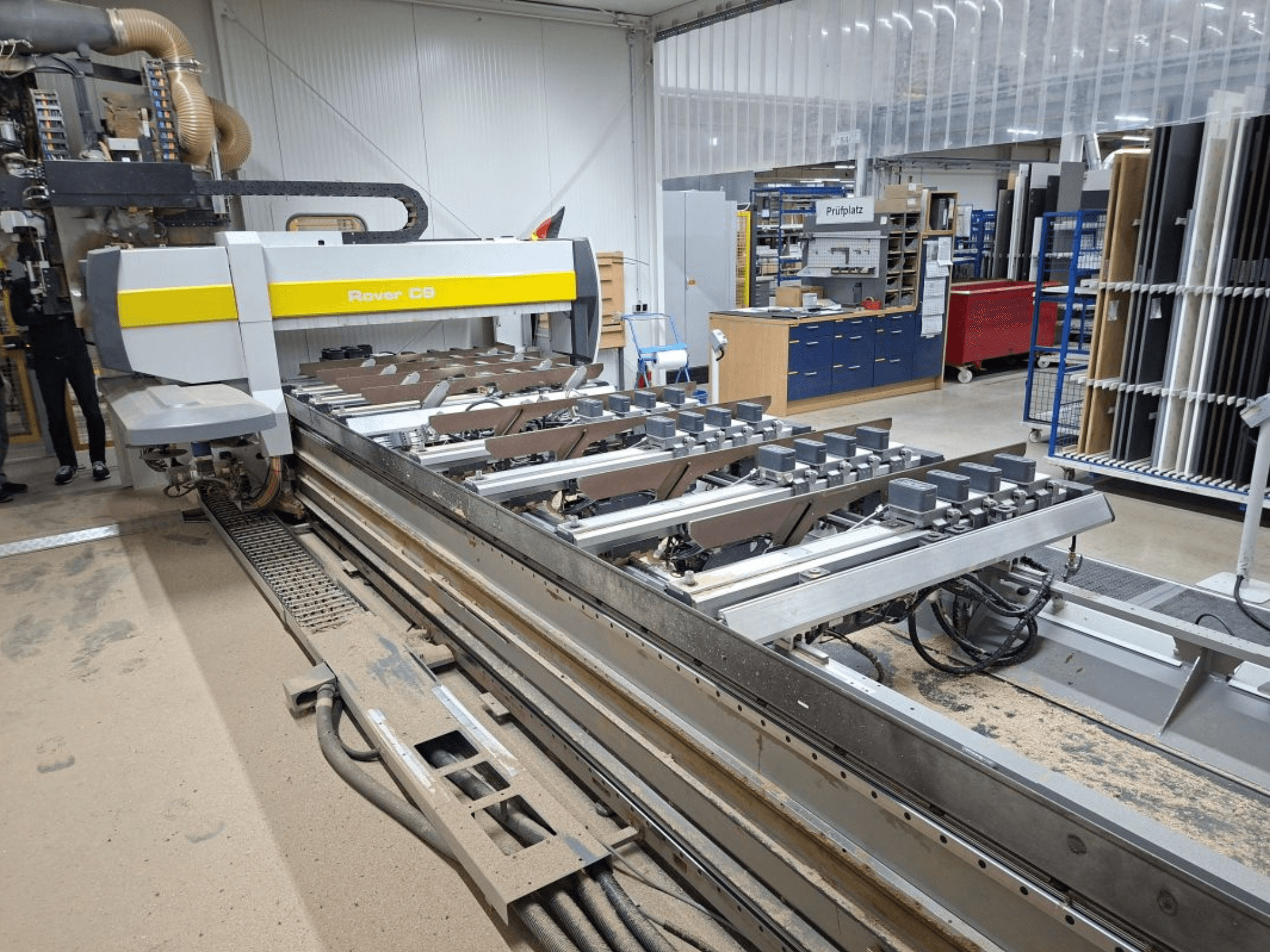 Stroj Rover C9 CNC v dílně; pohled zepředu ukazuje řezné nástroje a dopravní systém, na podlaze se hromadí prach.