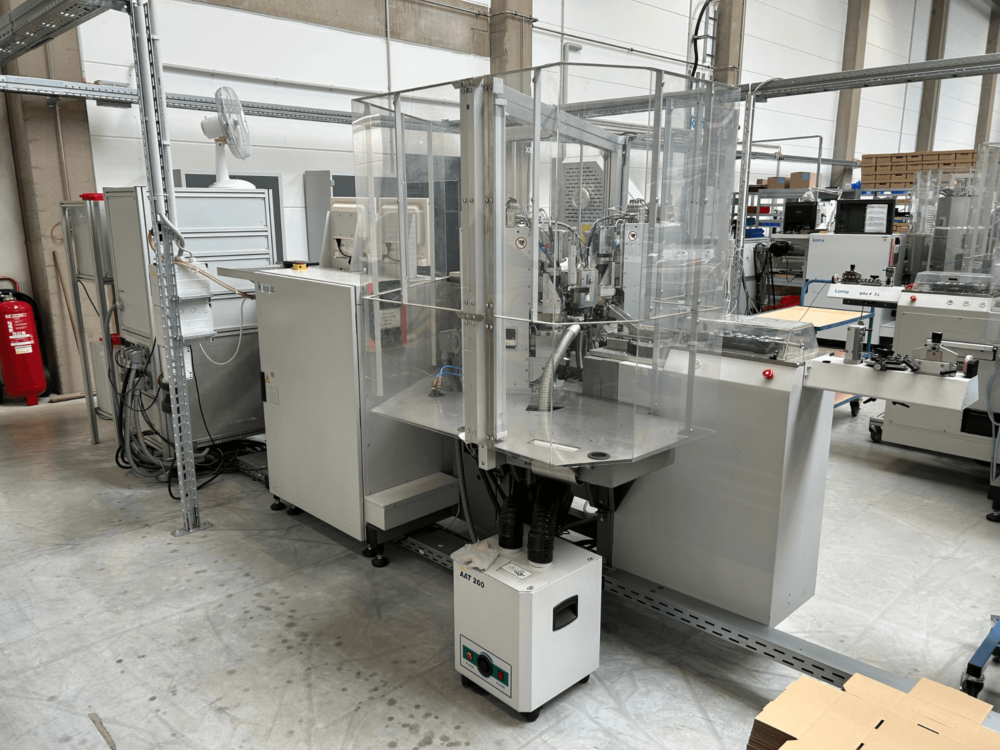 Automatizační zařízení KOMAX Alpha 433 L v továrním prostředí. Je vybaveno průhledným bezpečnostním krytem a různými ovládacími panely.