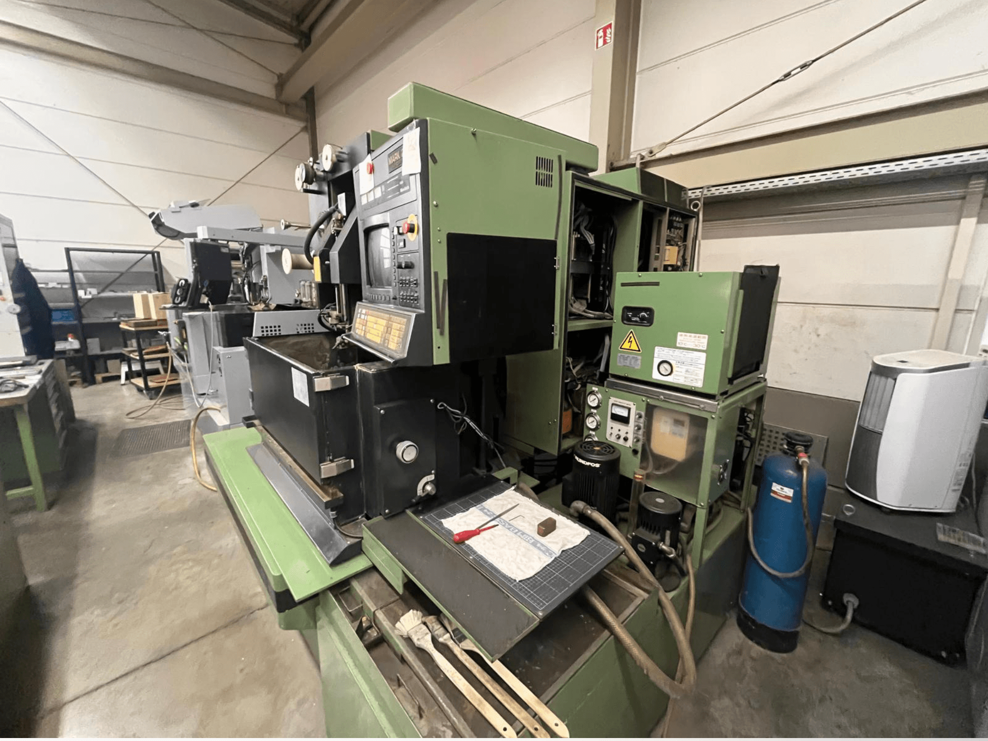 Zelený průmyslový stroj s ovládacím panelem, boční pohled; obsahuje hydraulické komponenty a pracovní plochu s nástroji.