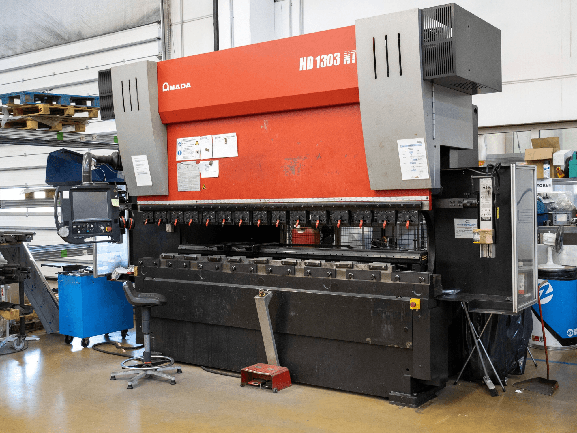 Hydraulická lisovací brzda Amada HD 1303 v dílně, pohled zepředu; obsahuje několik upínačů, ovládací panel a nastavitelnou stoličku.
