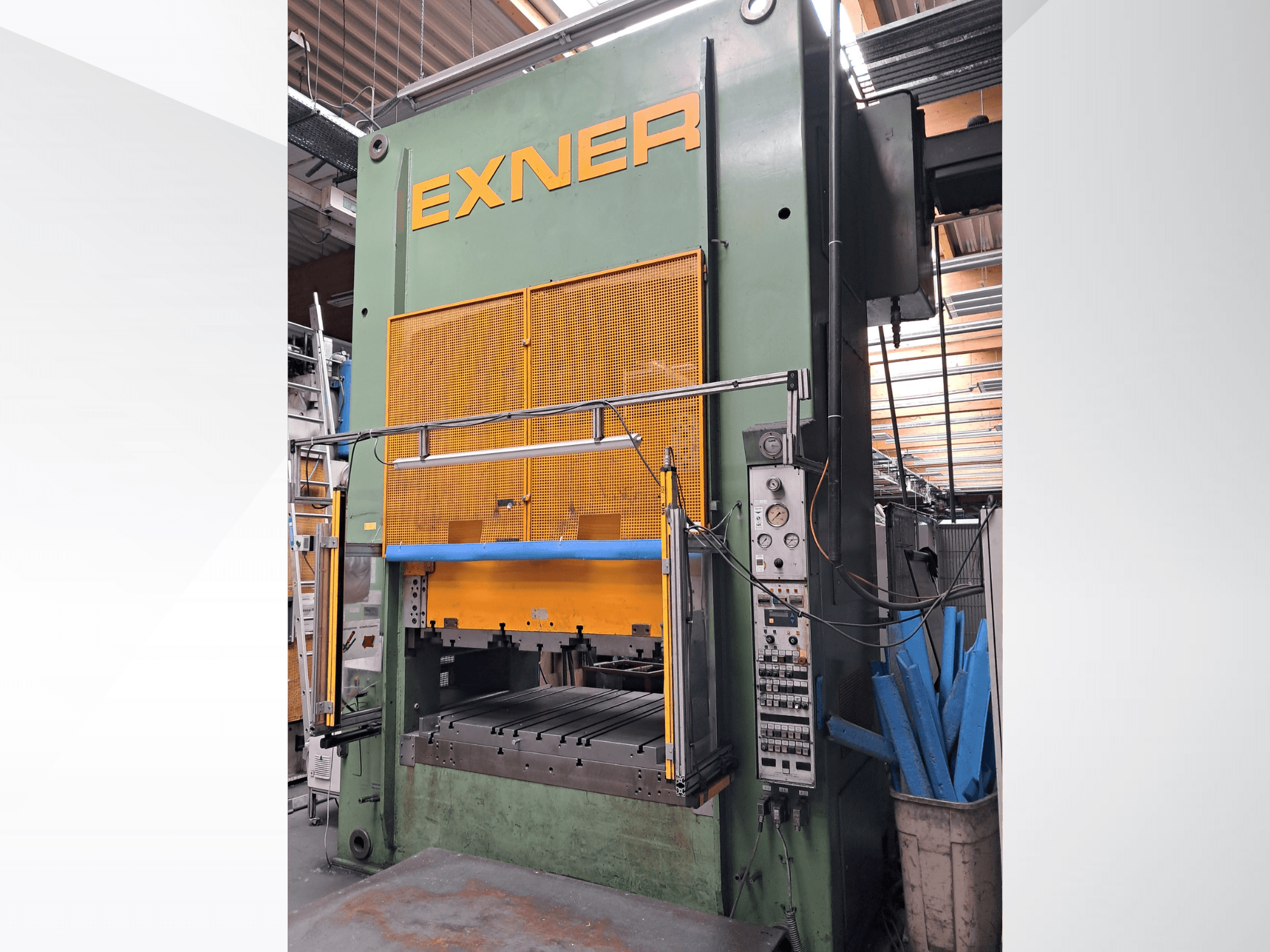 Hydraulický lis EXNER EX SBR 150, pohled zepředu; s ovládacími prvky, modrou bezpečnostní tyčí a ochrannou mřížkou, umístěný v dílenském prostředí.