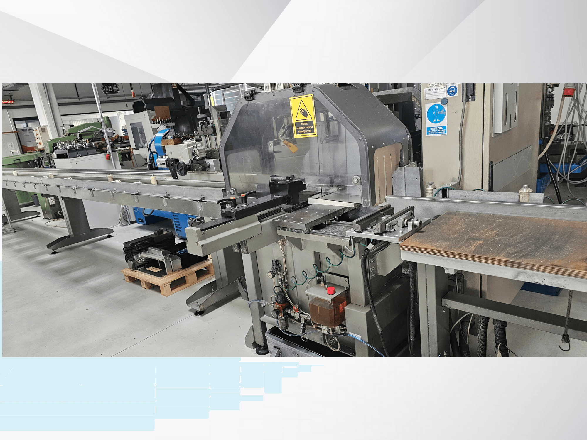 Průmyslový řezací stroj z bočního pohledu, s dlouhým dopravníkem, ovládacím panelem a bezpečnostními upozorněními.