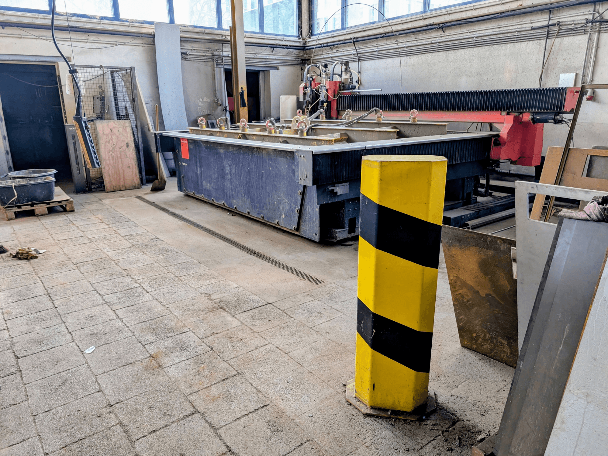 Stroj na řezání vodním paprskem Bystronic Byjet L 2030 v dílně, boční pohled, se žlutou bezpečnostní tyčí a okolními materiály.