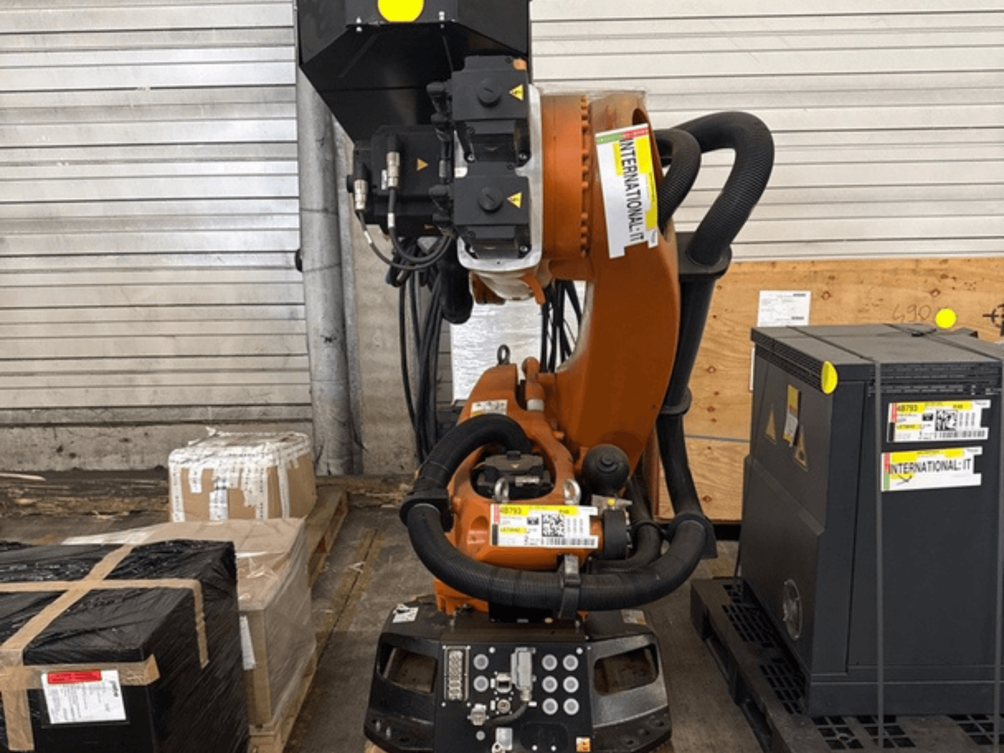 Robotické rameno KUKA KR 210 R2700 v čelním pohledu, vystavené na paletě vedle balíků a vybavení.