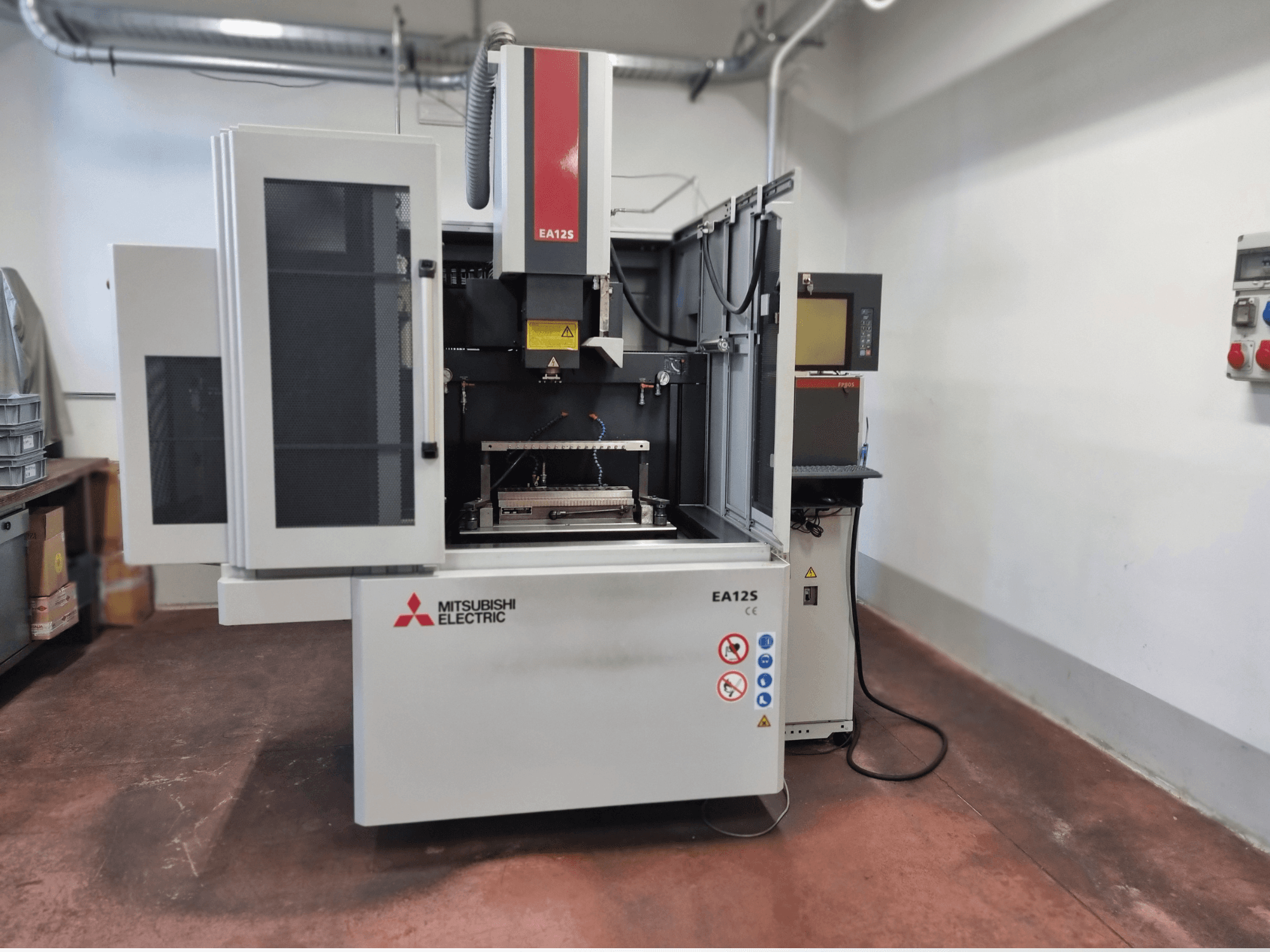 Drátový řezací stroj Mitsubishi Electric EA12S, pohled zepředu, otevřené dveře, ovládací panel a bezpečnostní štítky.