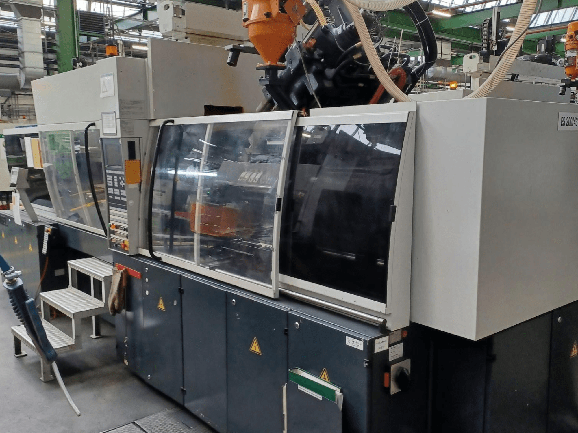 Pohled zepředu na vstřikovací stroj na plasty, značka a model neznámé, s průhlednými bezpečnostními dvířky a ovládacím panelem.