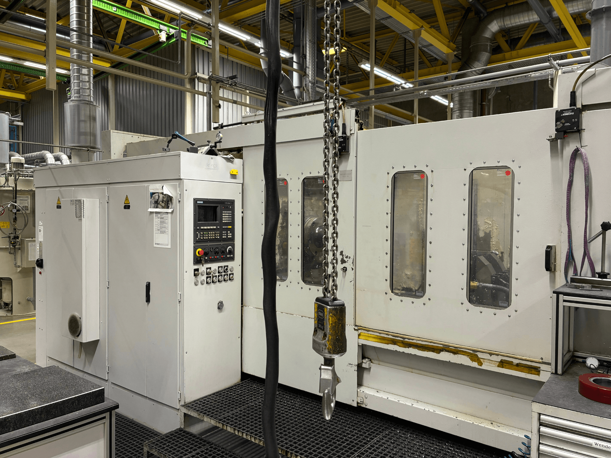 Průmyslový CNC stroj s ovládacím panelem, pohled z boku; s okny, řetězy a kovovými součástmi v továrním prostředí.