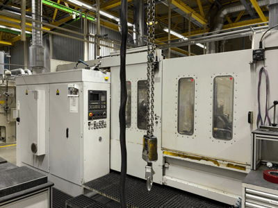 Průmyslový CNC stroj s ovládacím panelem, pohled z boku; s okny, řetězy a kovovými součástmi v továrním prostředí.